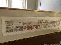 Long angled photo showing the full framed painting of houses with visible gold frame and white matting. Shows the panoramic format of the artwork.