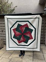 Full view of the handmade quilt held upright showing the geometric design, colors, textured quilting, and the tabs at the top for hanging.