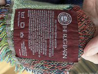 Close-up of the care and brand tag sewn into the throw blanket showing 100% cotton, made by The Rug Barn.