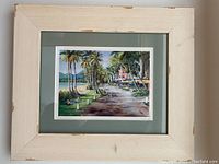 Overall front view of small framed painting with tropical scene and off-white wooden frame showing wear marks.