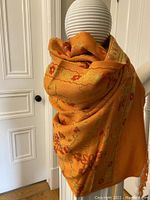 Shawl draped on a mannequin showing vibrant orange color with floral pattern and fringe edges.