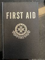 Front cover of vintage first aid book showing black textured hardcover with silver text and emblem.