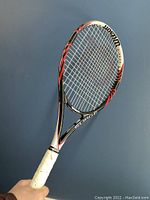 Side view of Wilson Surge BLK tennis racket showing black, white, and red design, white grip held in hand against blue wall backdrop.