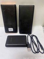 Three black power banks, two tall rectangular ones and one smaller rectangular one showing a wireless charging symbol, alongside one USB to micro-USB charging cable.