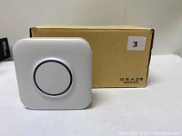 Photo of one white wireless doorbell receiver with a large circular button on the front, placed in front of a brown cardboard box.