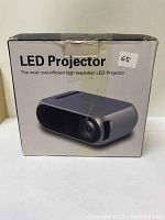 Photo of the retail box of a gray and black LED Projector with control buttons on top and lens on front, showing brand and basic product info