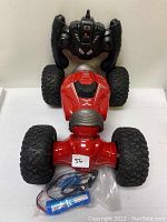 Photo shows red remote control car with large black wheels, black remote control, blue rechargeable battery, and charging cable all arranged together.