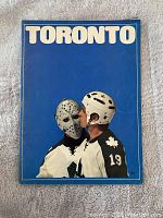Front cover of the 1970s Toronto Maple Leafs program, depicting a player wearing jersey 19 and helmet kissing a goalie mask against blue background.