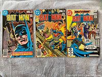 Front covers of three vintage Batman comic books laid out on a blanket surface.