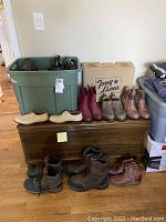 Wide shot showing multiple pairs of boots, shoes, scarves, hats on wooden chest and plastic storage bins. Shoes include beige sneakers, brown and red boots, hiking boots, dress shoes, scarves folded on top of plastic container.