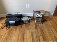 Four audio electronic devices arranged on hardwood floor against wall: Sony Mega Bass Boom Box CFS-D20, Philips Magnavox AZ1005 boombox, Emerson CD player, Sony CMT-NE3 mini stereo system with speakers and remote.