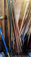 Multiple wooden handled garden tools including rakes, shovels, and post diggers leaning against wooden shed wall.