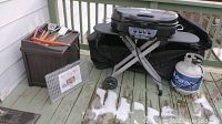 Front and side view showing Coleman gas grill, propane tank, grill utensils on hose storage container, and metal grill liner on deck