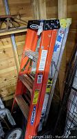 Two painters' ladders stacked together inside a wooden shed. One is orange with labels and safety instructions visible; the other is metallic with paint stains.