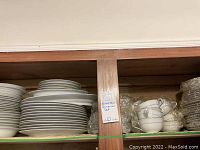 Stacked dinner plates, bowls, and platters on a shelf in a wooden cabinet. Some wrapped teacups and saucers are visible on the right side on the same shelf.