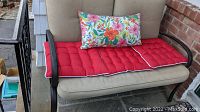 Front view of metal patio bench with beige seat and back cushions, red seat cushion with white piping, and colorful floral throw pillow. Partial view of nearby black metal railing and brick wall.