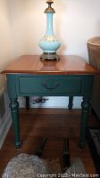 Side view of Ethan Allen wooden side table with wood stained top and painted green drawer and legs, topped with decorative lamp.
