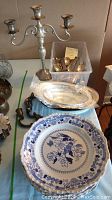 Photo of blue and white china plates with floral and bird pattern, three-arm silver-tone candelabra, and a plastic bin containing silver-plated flatware