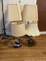 Photo showing two antique decorative electric table lamps with different fabric lamp shades and figurine bases placed on floor.