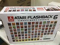 Shows the front of the box with Atari Flashback 6 branding and images of the 100 built-in games included on the console.