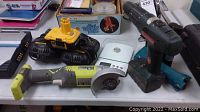 Overall view of lot items on table showing two drills, Ryobi angle grinder, chargers, and white kitchen scale.