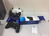 Four PlayStation controllers (two black, one blue, one white) and a boxed Qumo dual charging dock on table.