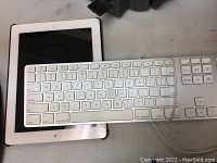 Shows front of wired Apple keyboard laying on top of tablet device with black screen and white bezel with home button.