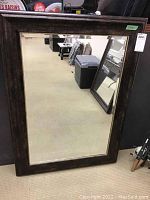Full view of the heavy framed mirror standing on the floor against a wall, showing the dark wide wooden frame and clear mirror glass reflecting room background.