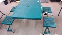 Green rectangular folding picnic table with four folding chairs open and set up on floor.
