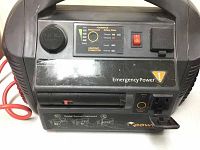 Front view of the Powerpack 300 Emergency Power Pack black casing with control panel and outlets visible. Red jumper cables placed on side.