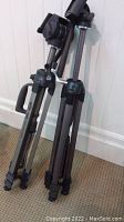 Two folded Optex T465 tripods standing against a wall on carpeted floor, showing black handles, adjustable parts and three legs each.