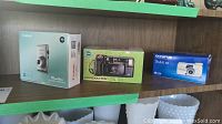 Original boxes for Canon PowerShot ELPH 300 HS, Minolta Freedom 202, Olympus Stylus 105 cameras on shelf