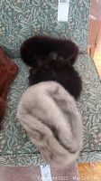 Three vintage fur hats displayed on a patterned green chair; visible are a cream/light mink hat, a dark brown fox hat, and a dark brown fur hat.