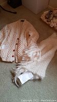Full view of cream fisherman's knit cardigan with matching cream wool shawl laid on carpet floor.