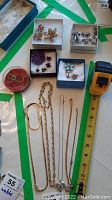Full view of jewellery lot arranged showing chains, lockets, earring and brooch sets, and the gold plated glove holder next to a tape measure for size reference.