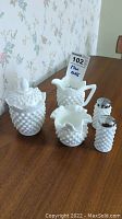 Full set of hobnail milk glass items including creamer, sugar bowl with lid, salt and pepper shakers, and small vase displayed on wood surface with floral wallpaper background.