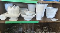 Shelf display of multiple milk glass vases of various shapes and sizes, including pedestal and hobnail designs.