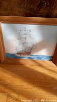 Framed canvas painting showing a sailing ship with full sails on blue water, light wood frame.