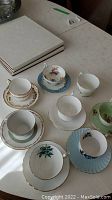 Seven assorted fine bone china tea cups and saucers on a white table.