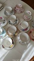 Photo showing twelve bone china tea cups and saucers on a table displaying floral designs and gold trim.