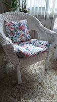 Front angle showing white wicker chair with floral cushions on seat and backrest, floral pattern includes pink, red, blue and green on white.