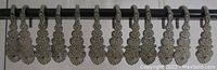 Front-facing image of 12 antique curtain hooks with ornate leaf motifs hanging in a row on a rod. Metal shows dark oxidation and wear.