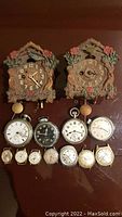 Two vintage Lux Keebler pendulette wall clocks with animated bluebird, surrounded by 11 assorted pocket and wrist watches displayed on a flat surface