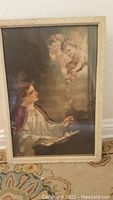 Tall vintage frame with artwork depicting woman and cherubs, glass covered, placed on carpet