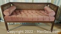 Full view of dusty rose velour upholstered bench with tufted cushion, wicker cane sides, and two rolled pillows