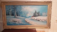 Full view of the framed winter landscape painting showing snowy scene with birch trees, fence, red cabin, and mountains