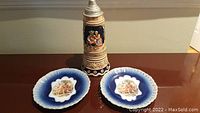 Photo shows two cobalt blue Royal Bavarian PMB China decorative plates flanking a tall German beer stein music box with painted scenes and a metal lid, all displayed on a wooden surface.