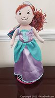 Front view of Groovy Girls princess doll wearing purple and turquoise dress with tag attached, standing on doll stand.