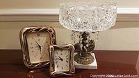 Photo showing the vintage Italian-style clear cut glass pedestal fruit bowl with cherub riding a fish on a marble base and dangling crystals beside two Italian silver table clocks.