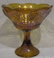 Front view of a tall round iridescent amber carnival glass compote with grape and leaf pattern on stem and bowl.